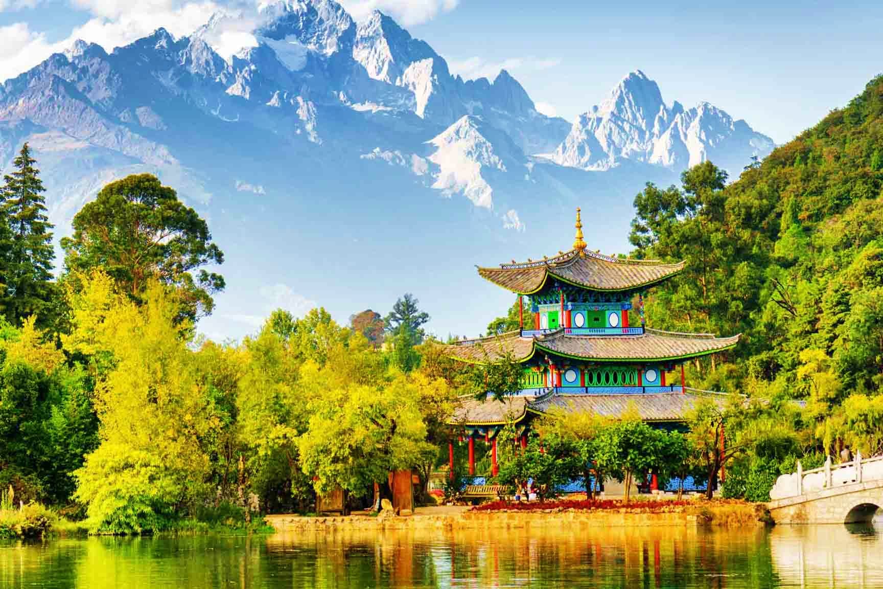 Traditional Chinese pagoda with upturned eaves reflected in river, surrounded by lush green trees with snow-capped mountains 